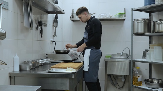 A professional chef prepares a gastronomic dish in a frying pan using ingredients and kitchen utensils. The chef is cooking in the kitchen of the restaurant. Cook's work in the kitchen.