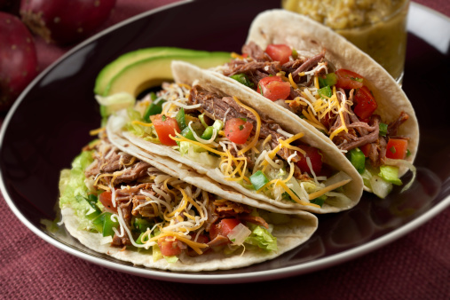 Three beef barbacoa tacos in soft tortillas.  Made with spicy shedded beef, lettuce, tomatoe, onion and cheddar cheese and served with a green salsa and avocado slices.