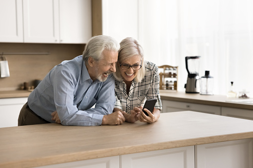 Happy adorable senior couple watching family pictures, online media content on mobile phone, using smartphone at kitchen table, laughing, feeling joy, talking on video call together