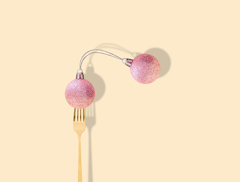 Fancy pink Christmas baubles connected and pricked on a fork on pastel beige background. New Year party background. Minimal style. Creative Christmas or New Year concept. Winter holidays idea.