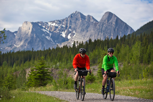 A man and woman go for a gravel road bike ride in the Rocky Mountains of Alberta, Canada. Gravel bicycles are similar to road bikes but have sturdy wheels and tires for riding on rough terrain. They both have a handlebar bag to carry food and bicycle tools.