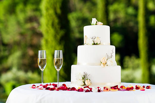 Wedding Cake Decorated With Flowers And Champagne Flutes On Table White wedding cake decorated with flowers and champagne flutes on table at reception in garden. Horizontal shot. 3 tiered white wedding cake stock pictures, royalty-free photos & images