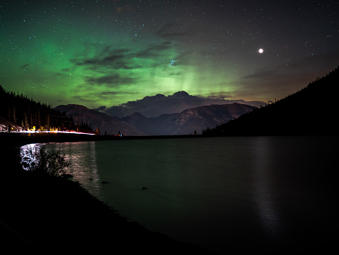 The Aurora Borealis illuminates the night sky above a mountain and lake with light streaks from a car passing above Canmore Alberta, Canada in the Canadian Rockies.