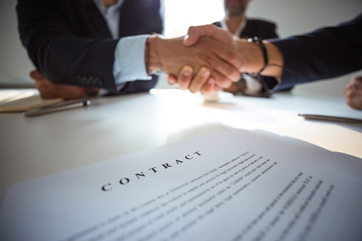 Two businessmen wearing suits sitting at the table in the  office and shaking hands over contract.  Close up of hands, unrecognizable people.