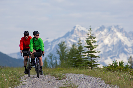 A man and woman go for a gravel road bike ride in the Rocky Mountains of Canada on a summer day. Gravel bicycles are similar to road bikes but have sturdy wheels and tires for riding on rough terrain. They both have a handlebar bag to carry food and bicycle tools.