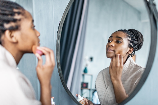 Young adult black woman in bathrobe applying concealer in the morning in bathroom