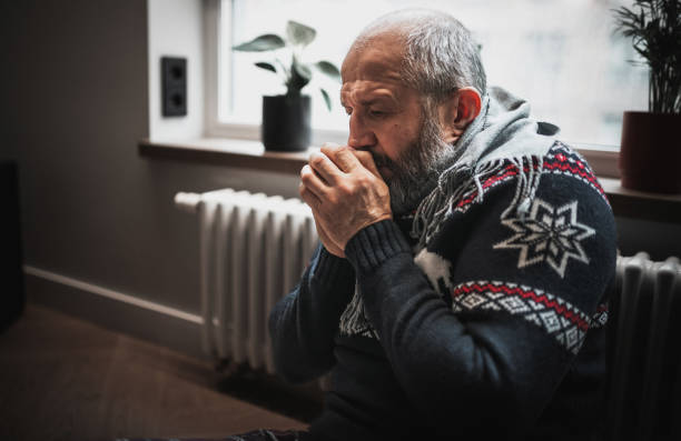 Unwell man feeling cold in home. He's trying to stay warm next to the radiator.
