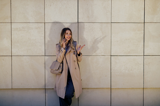 Smiling beautiful female wearing a trench coat leaning on a tiles wall and talking on a mobile phone. Woman making a phone call with a smartphone and enjoying talk with her friend.