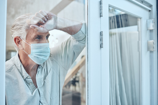 Lonely, face mask and senior man in the house for quarantine, protection and isolation during pandemic. Depression, frustrated and sad elderly guy with covid looking outside the window at his home.