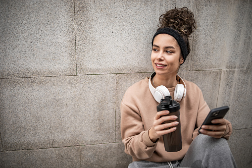 Young female athlete taking a break during outdoor exercise, sitting on stairs, drinking water and checking on her phone via activity meter how today's training went