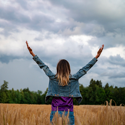 Rear view of a young woman celebrating life or practicing mindfulness standing in golden wheat field with her arms raised high. Under a cloudy summer sky.