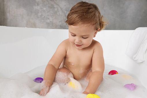 A happy baby is washing in a white bathtub with bathing toys. Funny child in the home bathroom. Kid aged about two years (one year nine months)