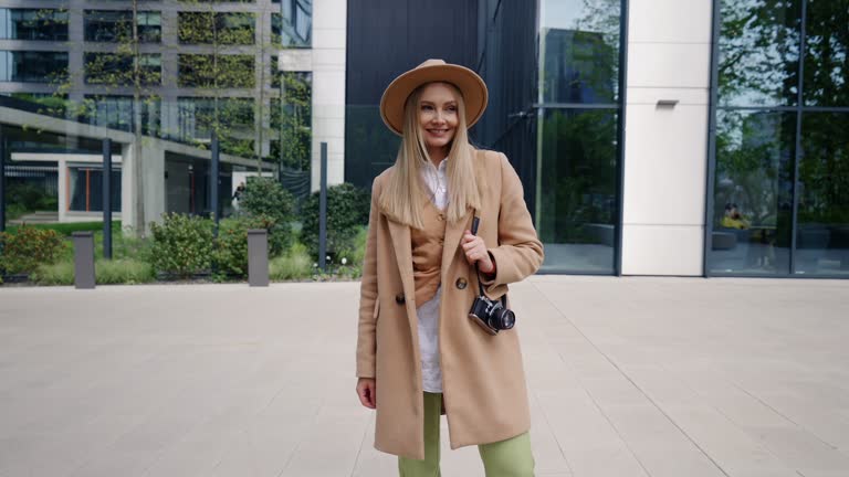 Stylish female photographer standing in modern city street. Beige coat and hat, retro style photo camera on her shoulder