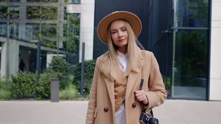 Stylish female photographer standing in modern city street. Beige coat and hat, retro style photo camera on her shoulder