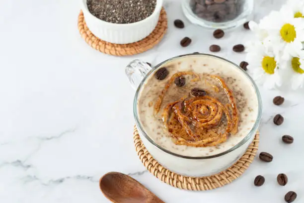 Chia seed pudding with coffee and peanut butter in glass, wooden spoon, and flowers on white marble table. Copy space. Healthy breakfast drink, clean eating vegan dessert.