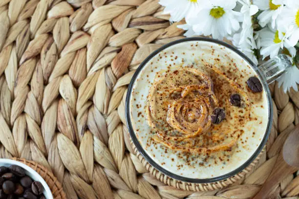 Creamy coffee cream chia seed pudding in glass on rustic rattan table with flowers and wooden spoon. Top view, copy space. Healthy clean-eating vegan breakfast or dessert.