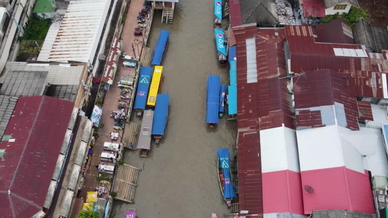 Boats pass through busy canal at Amphawa Floating Market. Tourists walk over bridge. Aerial view, dolly backwards.