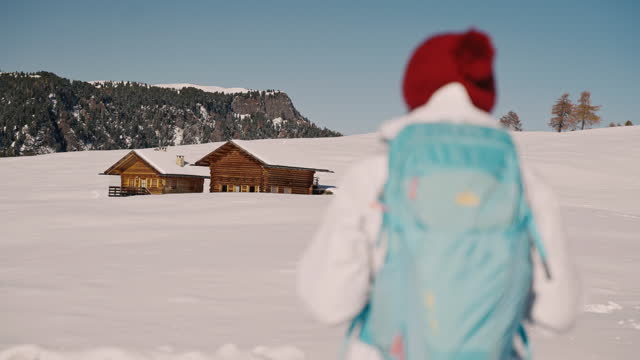 SLO MO Rear View of Young Female Backpacker With Wooden Lodges on Snow Covered Field in Dolomites