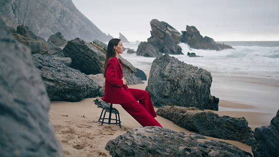 Woman sitting gloomy coast looking stormy ocean. Girl posing on chair at beach.