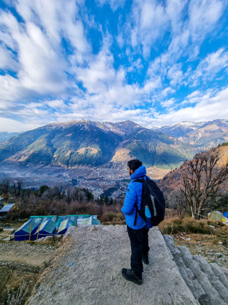 Male trekker hiker standing looking down at village houses and himalaya mountains in distance and cloudy sky in manali, kullu, shimla, kedarnath Male trekker hiker standing looking down at village houses and himalaya mountains in distance and cloudy sky in manali, kullu, shimla, kedarnath in India kullu stock pictures, royalty-free photos & images