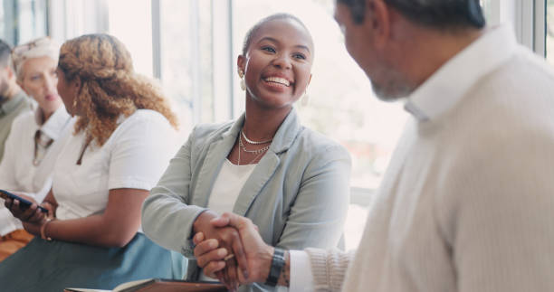 People, shaking hands and queue for meeting, smile and waiting room for onboarding, networking and interview. Black woman, man and handshake for hello, kindness or welcome for recruitment opportunity