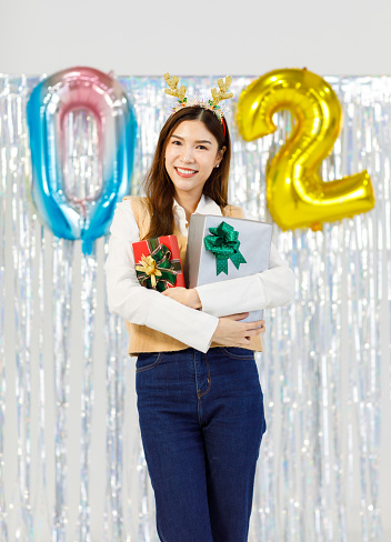 Portrait studio shot of Asian cheerful female model wearing reindeer antlers headband smiling look at camera holding wrapped presents gifts boxes celebrating New Year 2024 and Merry Christmas Eve.