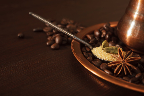 Closeup of cardamom pods, anise and brown sugar in a teaspoon with coffee beans and shiny copper plate and pot as a background.