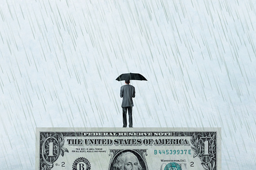 A man holds an umbrella to protect himself from the rain as he stands on top of a one dollar bill.