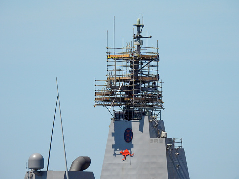 The main mast of HMAS Canberra, a Canberra Class Amphibious Assault Ship of the Royal Australian Navy, is surrounded by scaffolding.  She is docked at Garden Island naval base in Sydney Harbour.   This image was taken on a sunny afternoon on 11 November 2023.