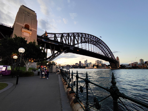 People walking on the waterfront at the iconic Sydney harbor bridge. It is the sixth longest spanning-arch bridge in the world and the tallest steel arch bridge, measuring 134 m from top to water level, on Sydney harbor, Australia.