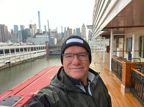 A man in a black woolen hat takes a selfie on the deck of a ship. In the background, we can see New York City.