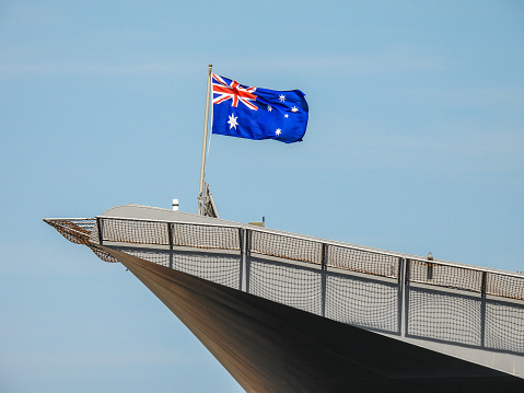 The Australian flag flies on the bow deck flagpole of HMAS Adelaide of the Royal Australian Navy docked at Garden Island, Sydney Harbour. Safety netting around the flight deck casts a shadow over the hull.  This image was taken on a windy and sunny afternoon on 11 November 2023.