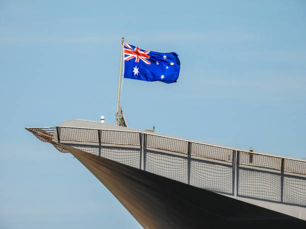 HMAS Adelaide Remembrance Day Flag The Australian flag flies on the bow deck flagpole of HMAS Adelaide of the Royal Australian Navy docked at Garden Island, Sydney Harbour. Safety netting around the flight deck casts a shadow over the hull.  This image was taken on a windy and sunny afternoon on 11 November 2023. australian navy stock pictures, royalty-free photos & images