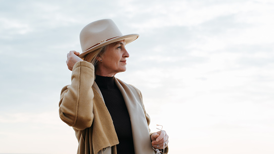 Smiling elegant mature lady in coat touching round brown hat standing against sky and looking away, copy space. Lonely senior woman outdoor lifestyle, mood.