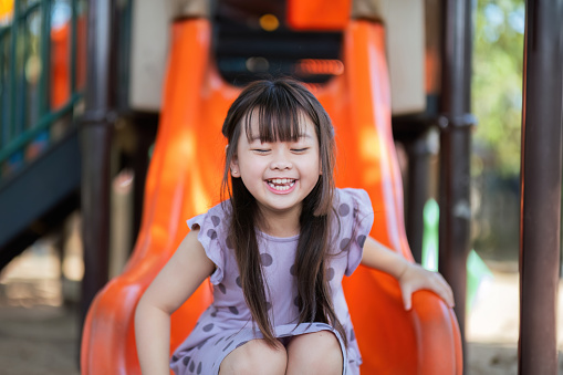 Smiling Asian little girl with fun sitting on slider at playground in the park. Education activity outdoor concept.