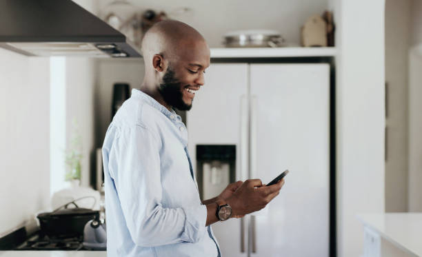 istock Man, smile and typing on smartphone in kitchen for social network, update multimedia subscription and chat at home. Happy african guy download mobile app, reading notification and search for contact 1798152517