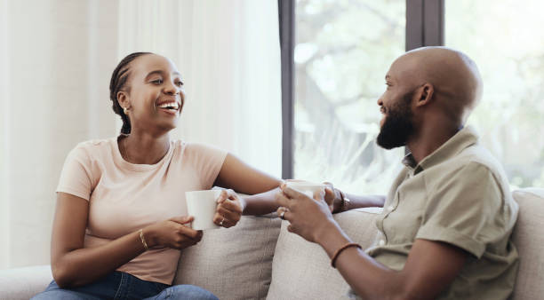 istock Couple, communication and smile on sofa with tea for relax, discussion and peace on weekend in living room. Black people, man and woman with conversation or talking on couch for coffee, love or care 1798152525