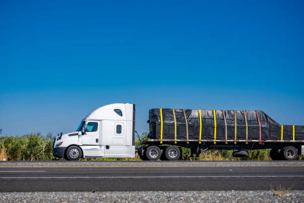 White high cab profile carrier big rig semi truck transporting cargo on flat bed semi trailer running on the one way highway road with olive gardens on the side going to distribution warehouse