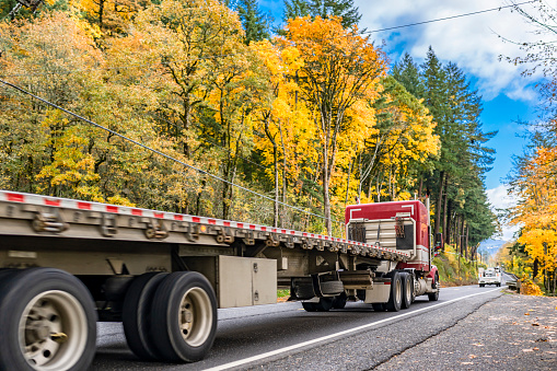 Red classic long hauler carrier big rig semi truck with extended cab transporting empty flat bed semi trailer running on the autumn narrow road with yellow trees on mountain hill in Columbia Gorge
