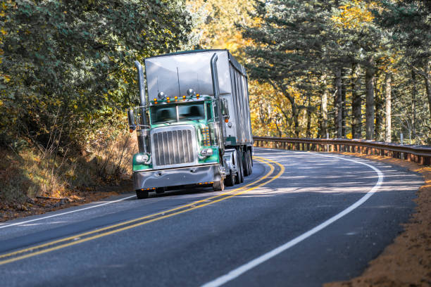 istock Classic green big rig semi truck tractor with day cab transporting covered bulk semi trailer driving on the narrow winding road in autumn forest 1798227221
