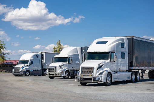 Industrial long hauler carriers white big rig semi trucks tractors with loaded semi trailers standing in row on the highway road rest area parking lot take a break for truck driver rest