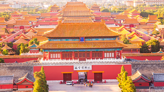A view on a big square inside a building complex of Forbidden City in Beijing, China. The buildings have very richly decorated rooftops, with elements of gold. Thera are lots of people on the square.