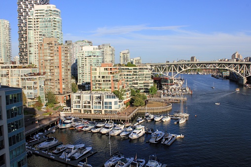Vancouver, Canada – July 19, 2023: A scenic view of a harbor featuring several moored boats and a bridge connecting two sides of the water, surrounded by a variety of buildings