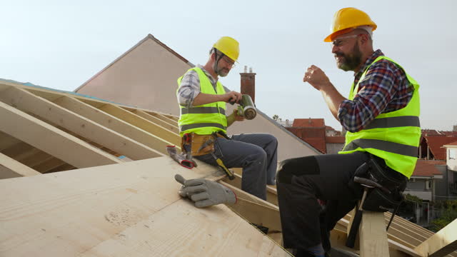 Mid-adult Caucasian male roofers taking a break from work, while sitting on a roof beam and drinking coffee
