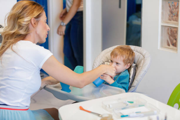 Feeding therapist assisting blond little boy in high chair, he is drinking from special cup