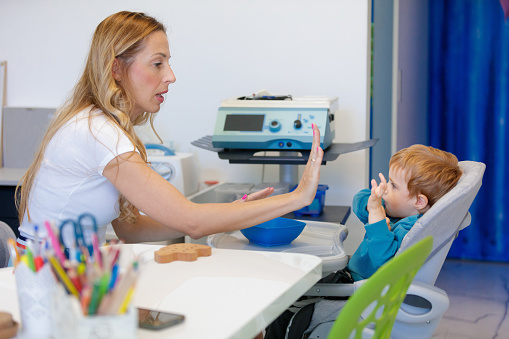 Blond toddler sitting in high chair and looking at feeding therapist, she is giving him high-five, oral motor skills, occupational therapy