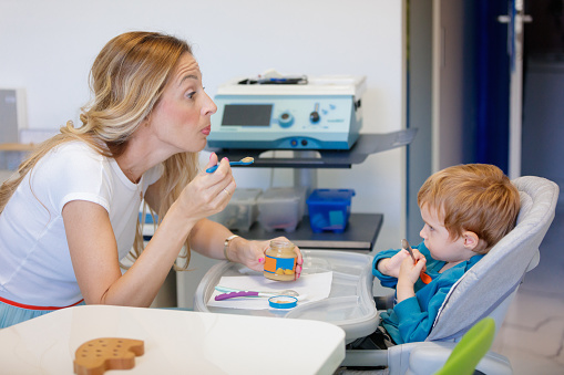 Feeding therapist holding baby food jar and pretending to eat with spoon, little boy in high chair is looking at her, oral motor skills, occupational therapy