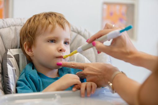 Blond toddler sitting in high chair and clamping mouth shut, looking at feeding therapist, she is touching his chin and lips, oral motor skills, occupational therapy