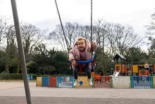 Full shot of a baby on a swing in a public play park with a jungle gym in the background. She is smiling, looking away from the camera. She is wearing warm clothing on a cold winter morning. The park is located in Gateshead.\n\nVideos are also available for this scenario.