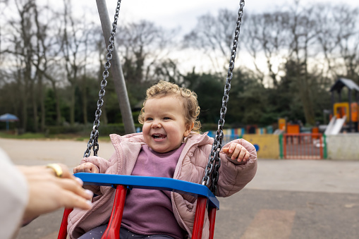 Waist-up portrait of a baby girl being pushed on a swing in a public play park. She is smiling, looking away from the camera at the person pushing her. She is wearing warm clothing on a cold winter morning. The park is located in Gateshead.\n\nVideos are also available for this scenario.